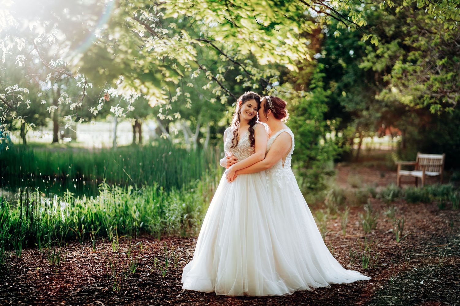 Two brides in white gowns embrace in a sunlit garden setting.