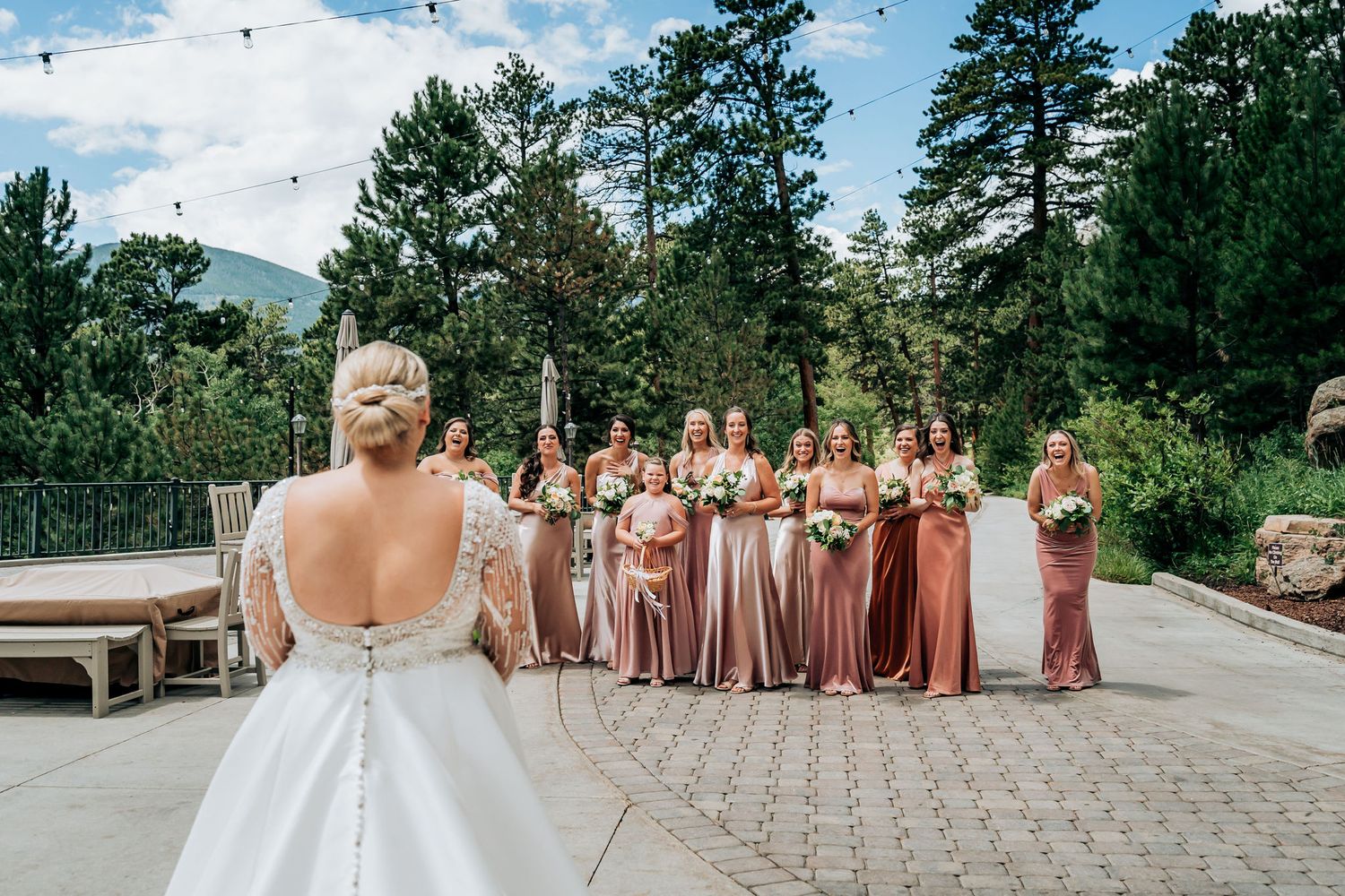 Bridesmaids in flowing earth-toned dresses stand behind a bride in a backless white gown at an outdoor venue.