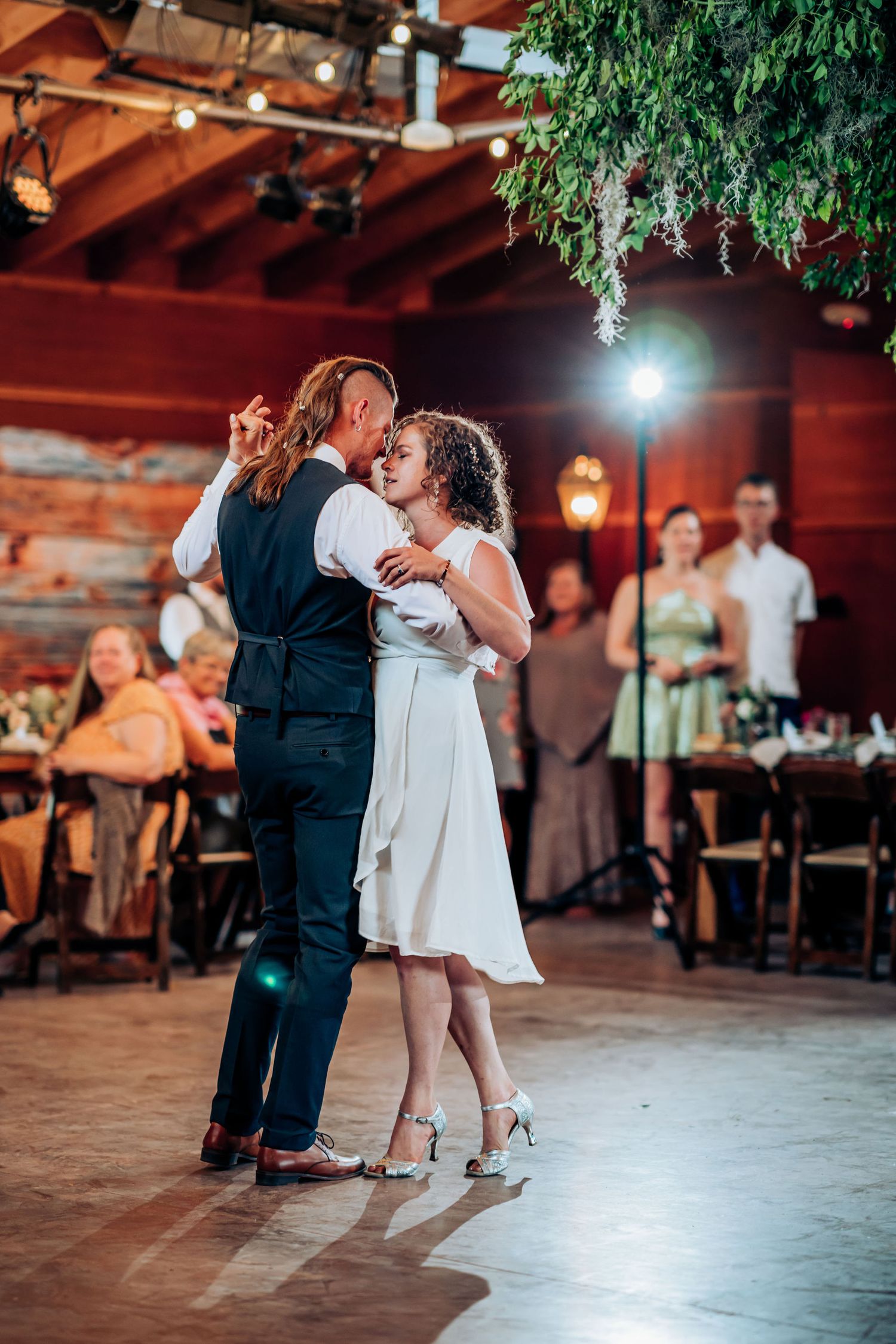 A couple dancing intimately at a wedding reception under hanging greenery and warm lighting.