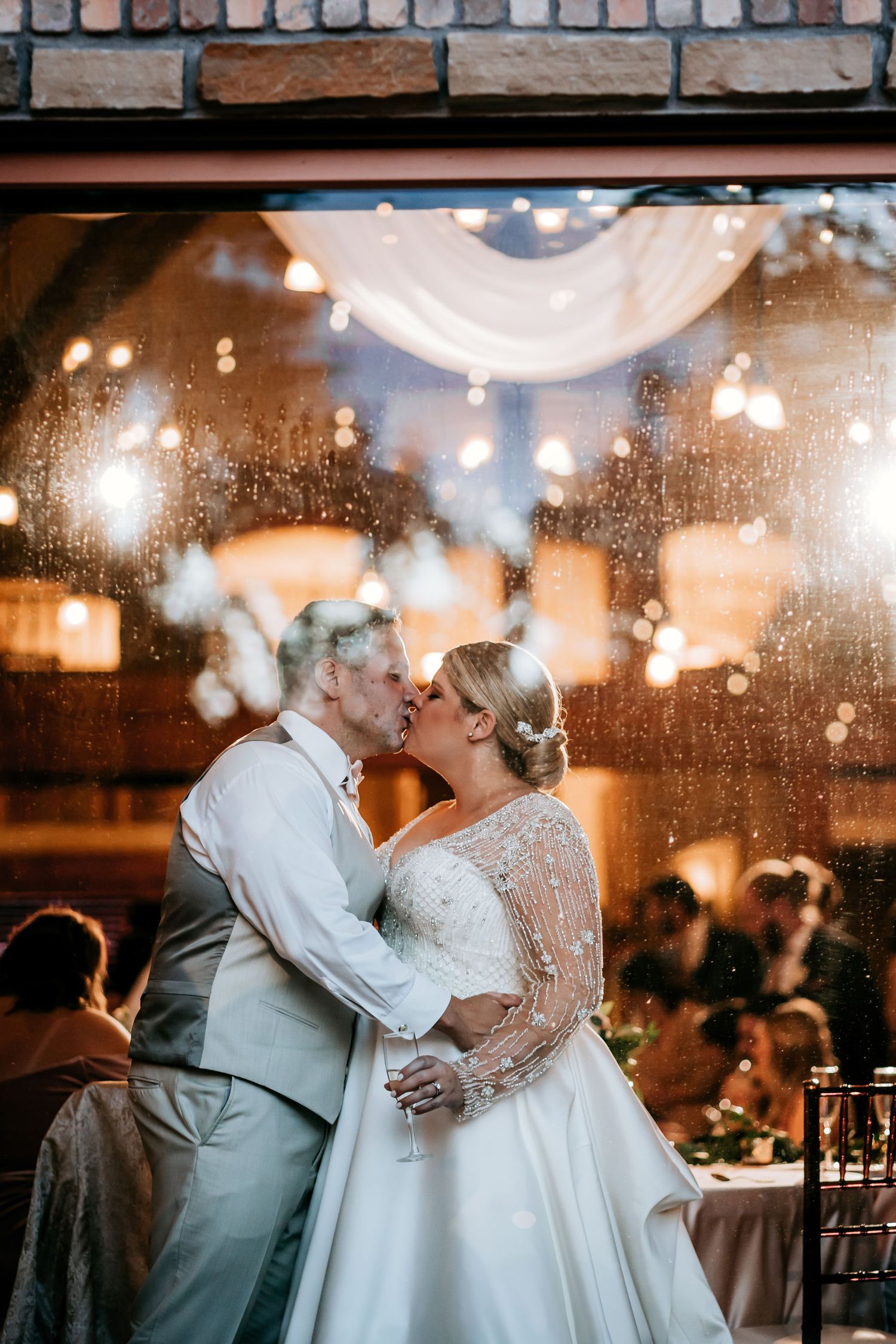 A romantic dance moment captured through rain-spotted window glass with warm lighting.