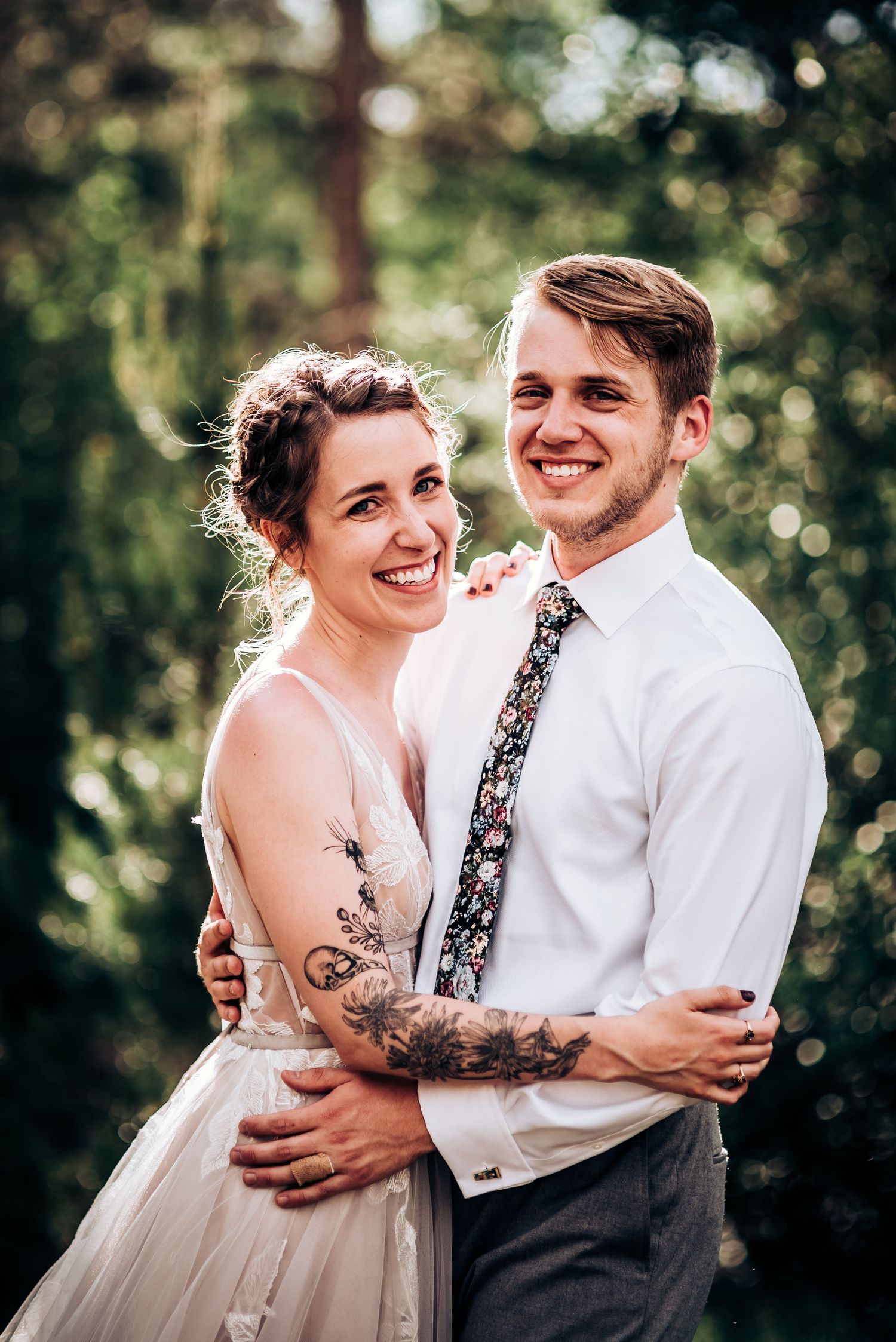 A wedding couple in a forest setting embraces and laughs together, with the bride wearing a tulle gown and showing arm tattoos.