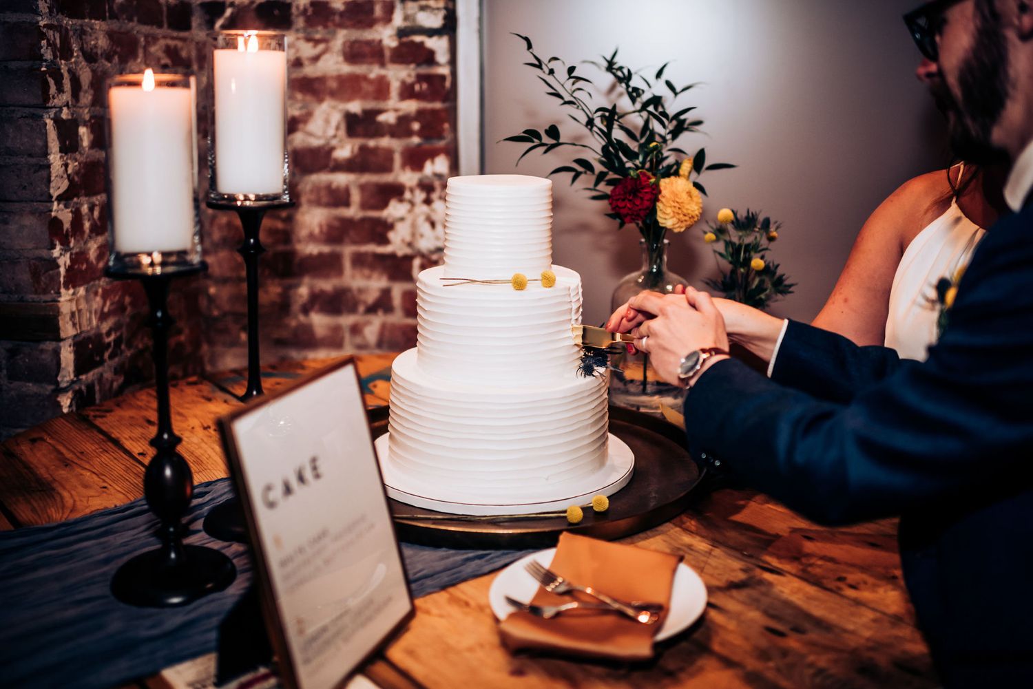 White tiered wedding cake with gold accents displayed on wooden table with candles and menu card.