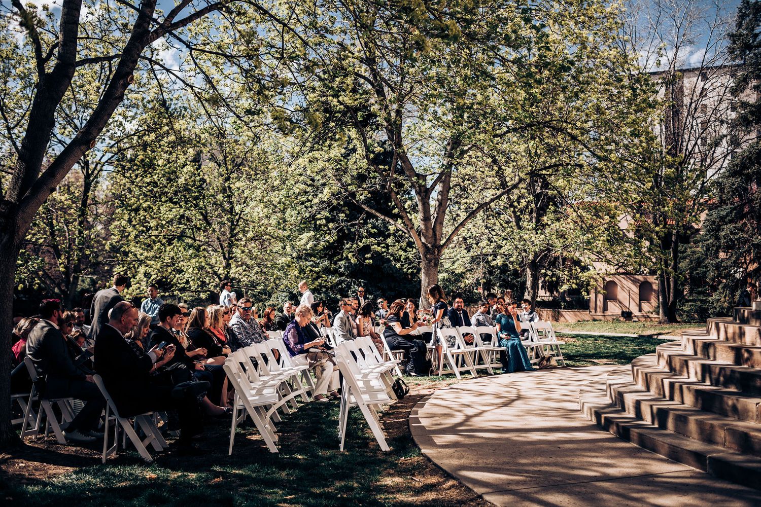 Guests seated in white chairs at an outdoor wedding ceremony with blooming trees in background.