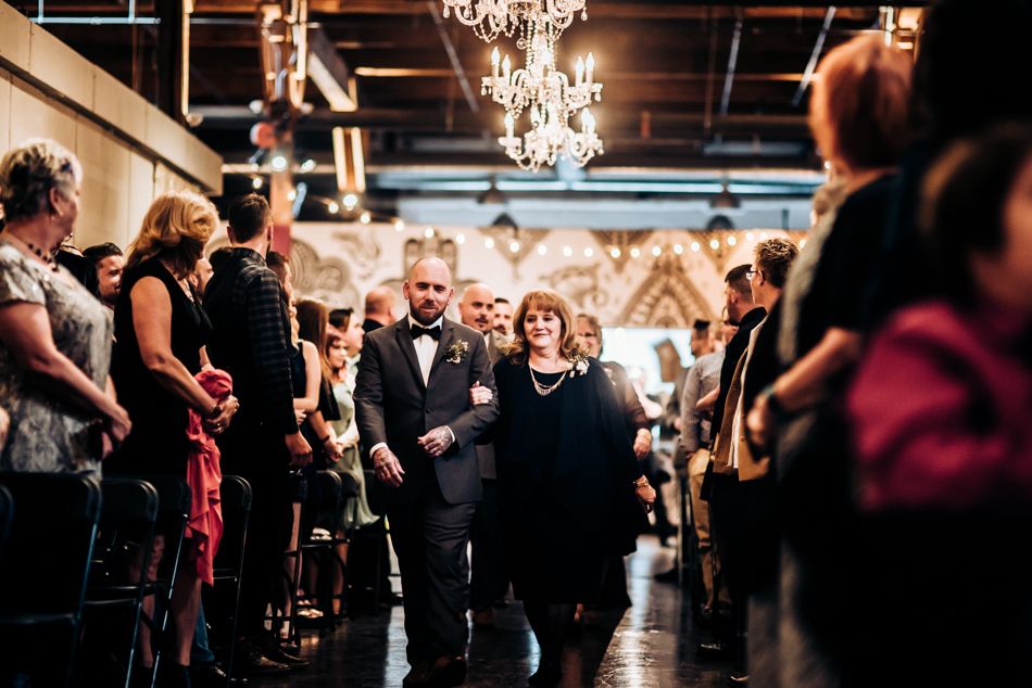 Wedding ceremony guests stand in a venue with industrial ceilings and elegant chandeliers.