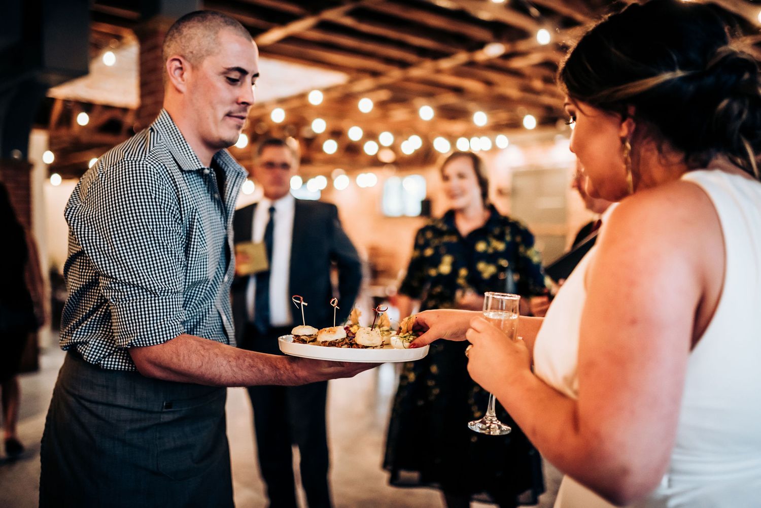 Server presenting appetizers to guests at an elegant indoor wedding reception with warm lighting.