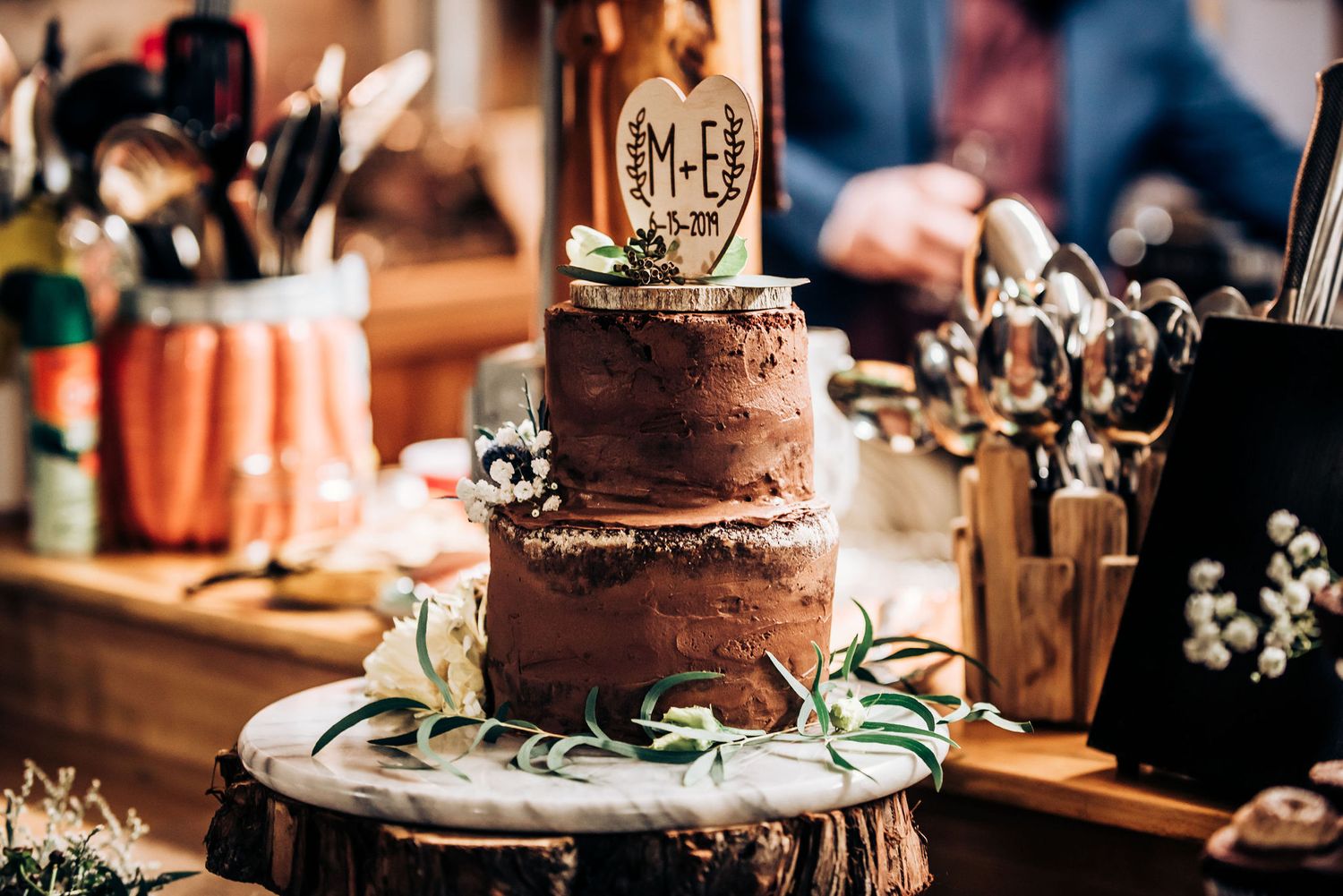 Rustic chocolate wedding cake decorated with greenery on a wooden cake stand.