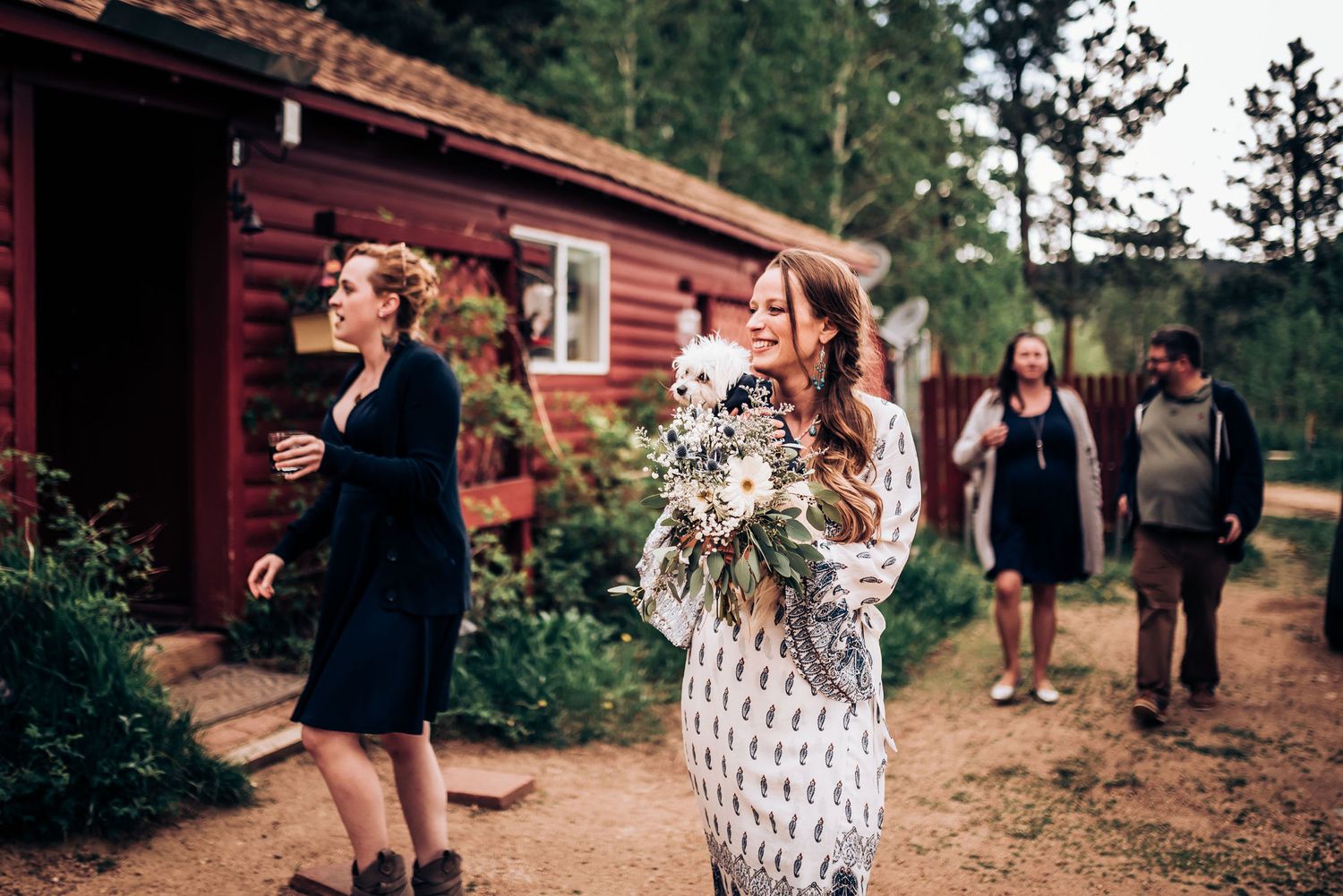 Wedding guests gather outside a rustic red cabin holding floral arrangements and drinks.