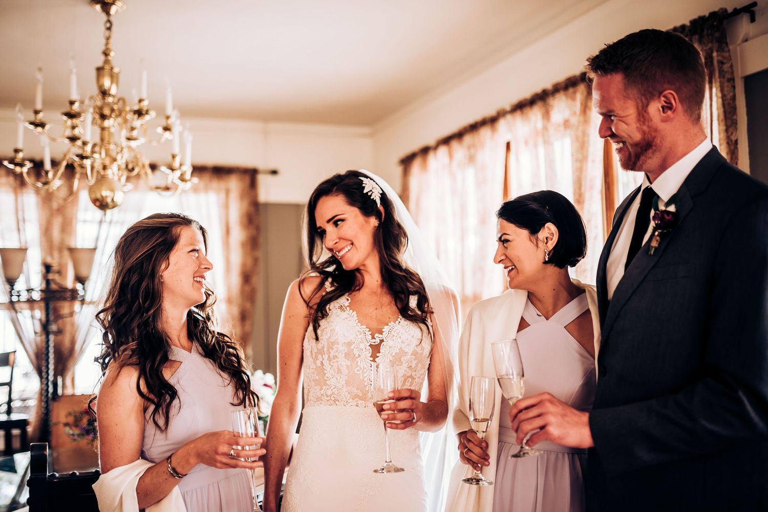 Elegant wedding party sharing champagne beneath a crystal chandelier in a brightly lit room.