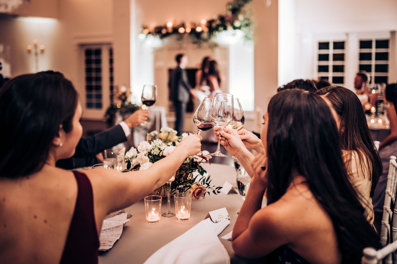 Wedding guests raise wine glasses in a toast during elegant indoor reception with soft lighting.