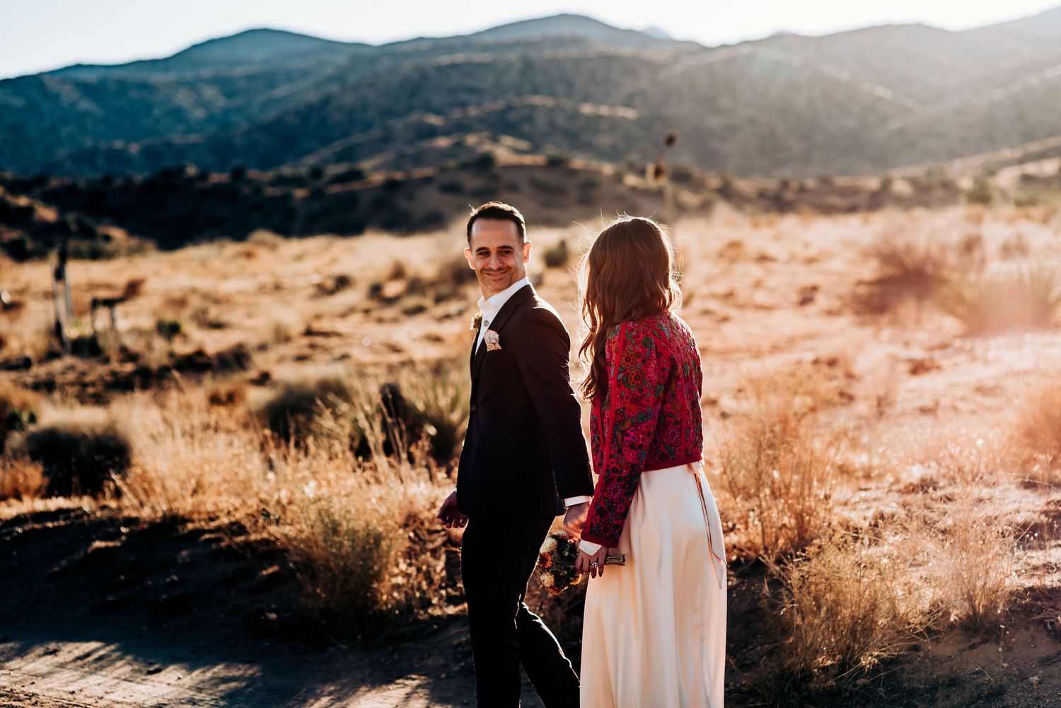 A couple in formal wear poses against a scenic desert mountain backdrop.