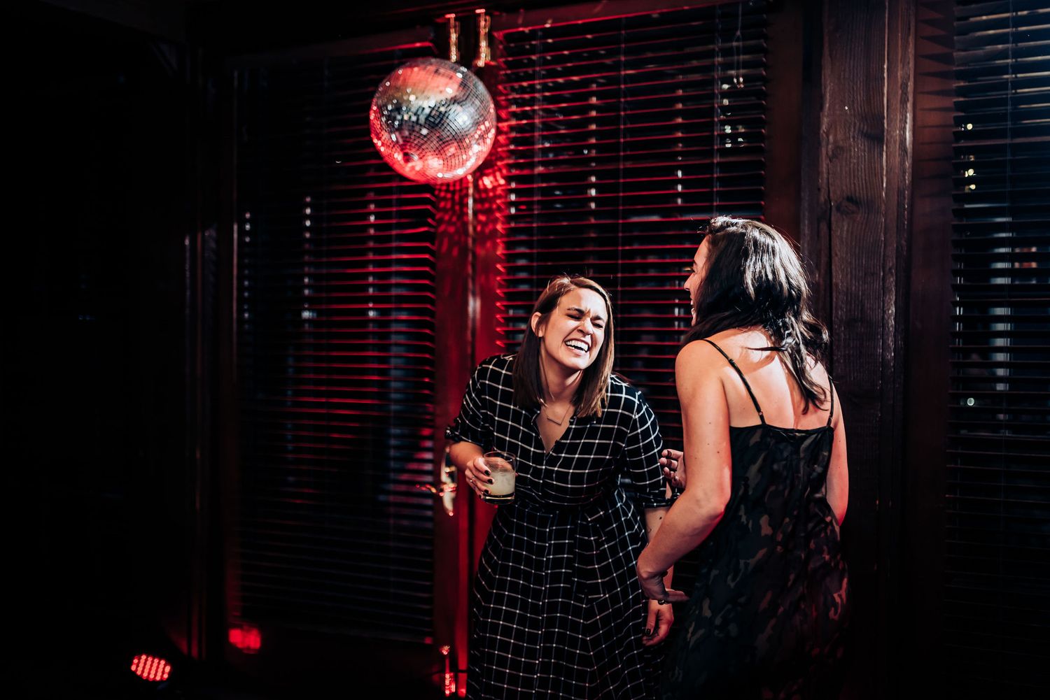 Two partygoers chatting beneath a glowing red disco ball in a dark room with venetian blinds.