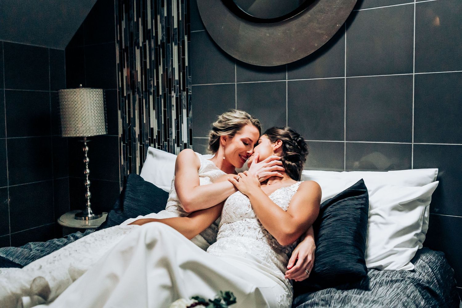 A romantic wedding couple share an intimate moment while sitting together with pillows on their bed.