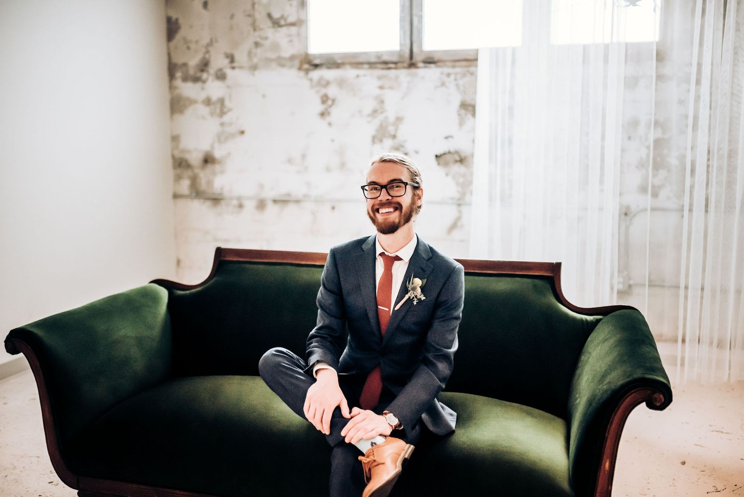 Candid portrait of groom seated on vintage green velvet sofa in industrial space.