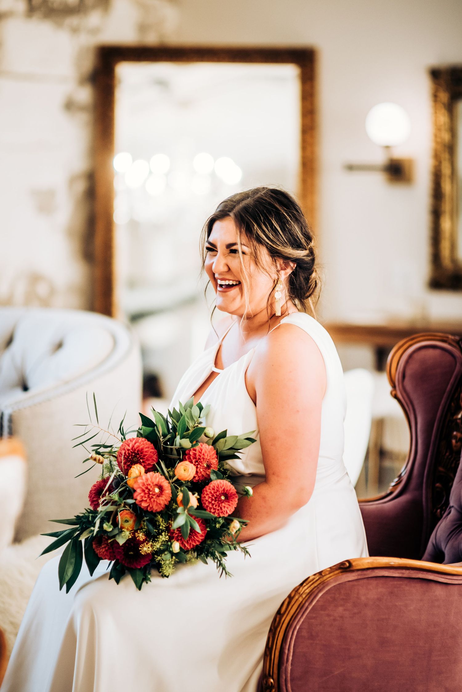 Person in elegant white dress laughs while holding colorful wedding bouquet in ornate room.