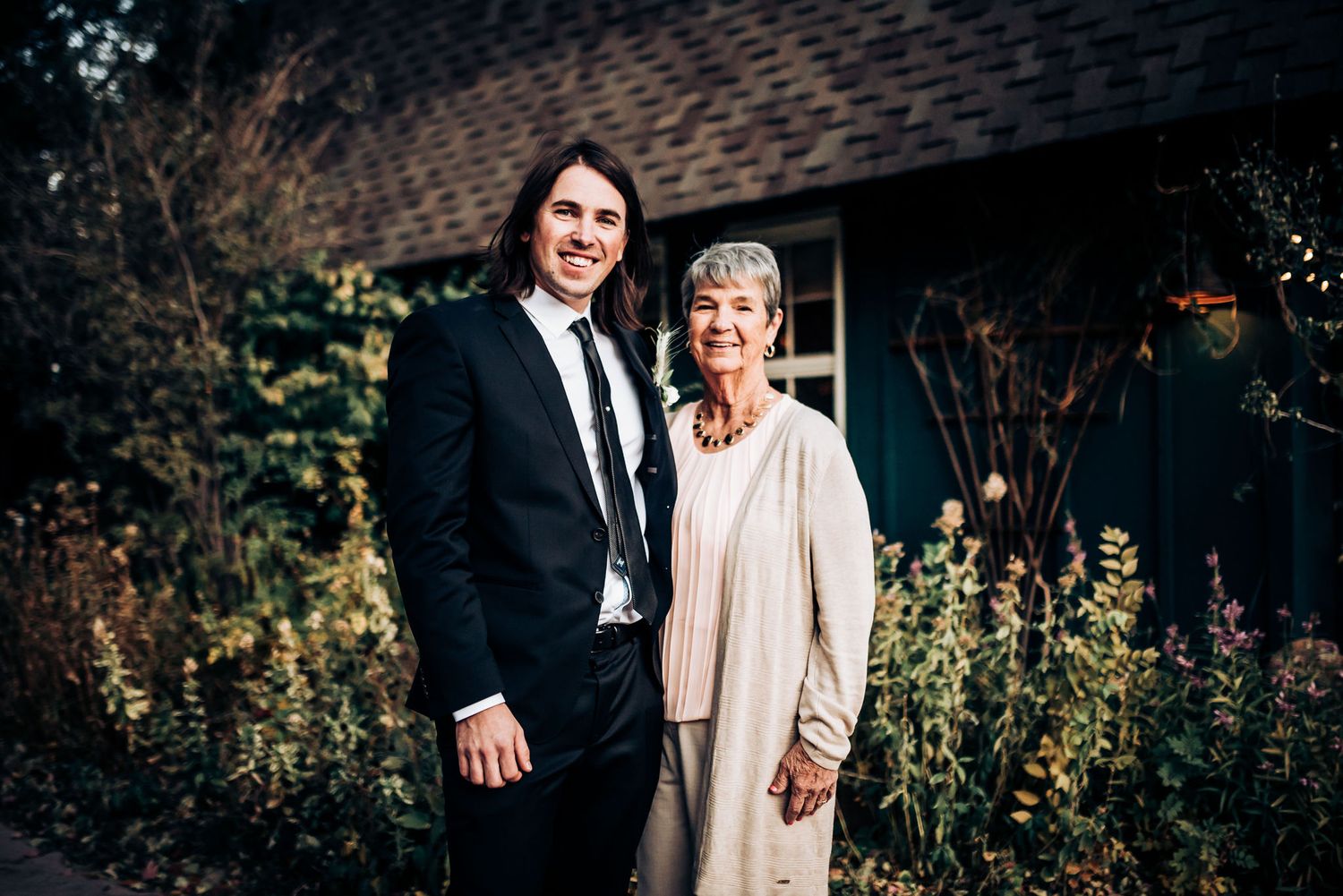 Formal portrait in garden setting with flowering bushes in background.