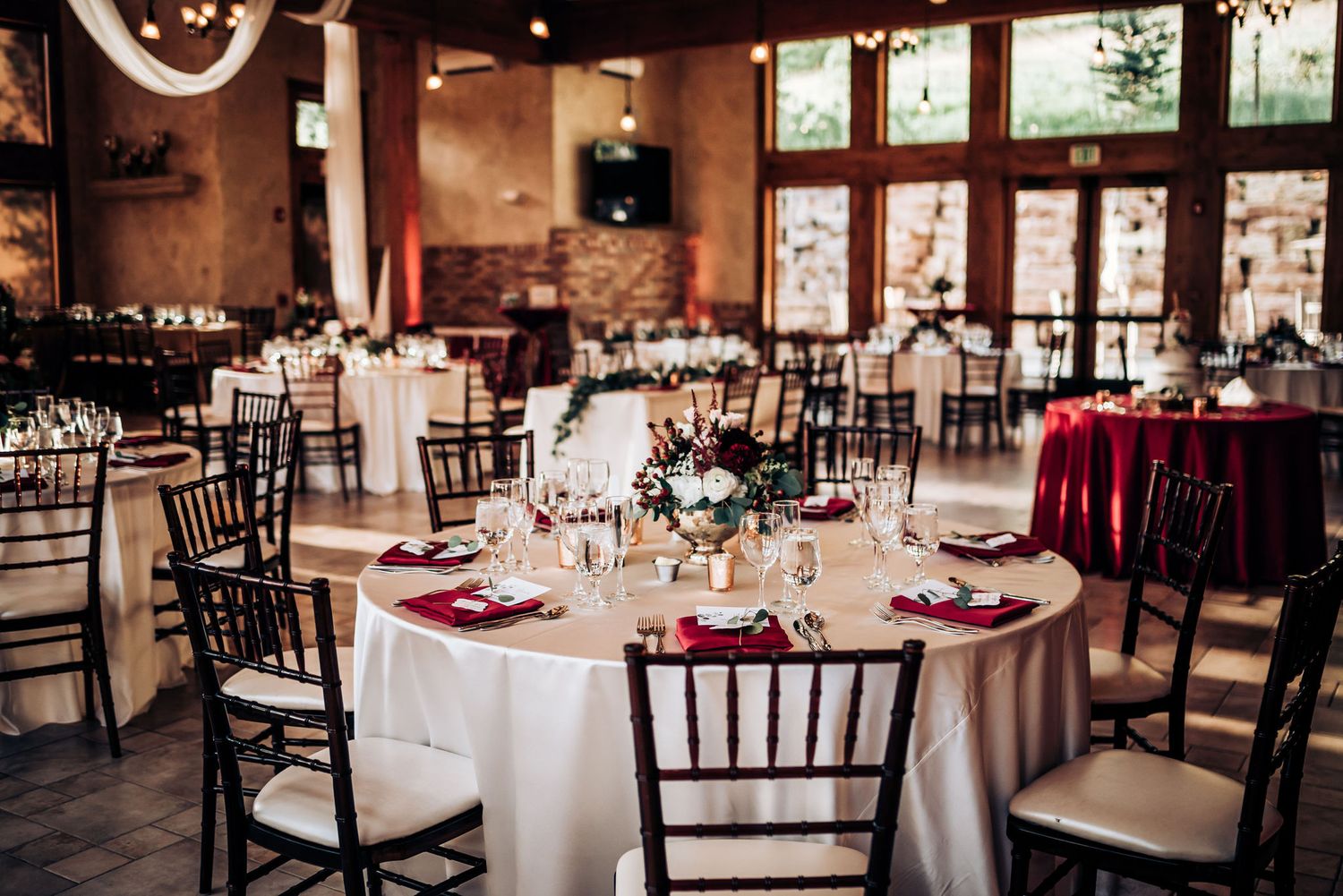 Wedding reception hall decorated with round tables, white linens and dark chiavari chairs in natural lighting.