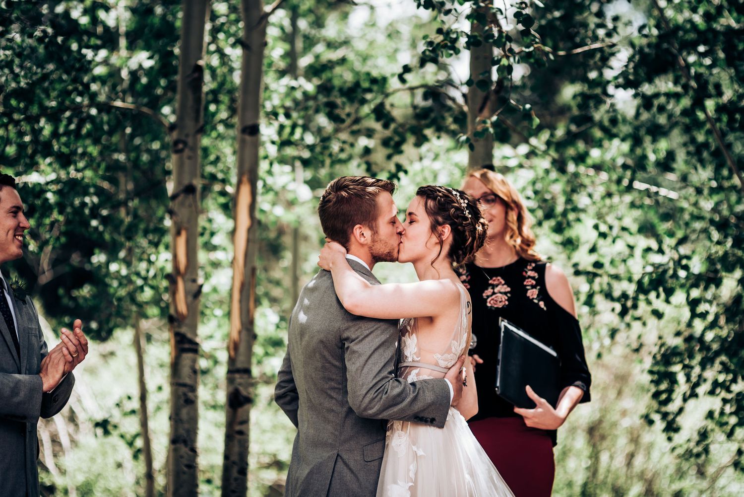 Newlyweds share their first kiss during a forest wedding ceremony as guests applaud.