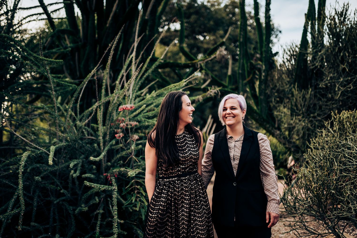 Two people share laughter while standing among tropical plants and cacti.