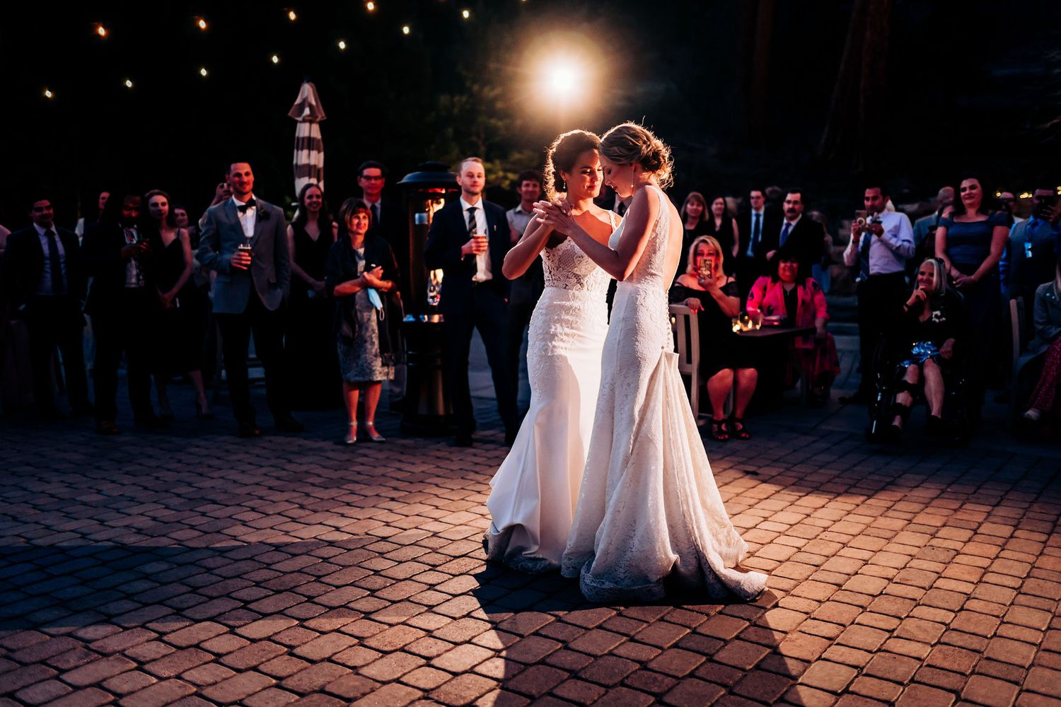 Couple in matching white wedding dresses dance together surrounded by guests on a paved courtyard.