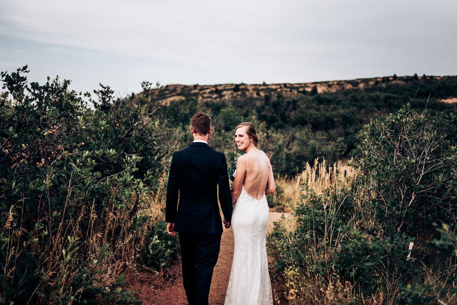 A couple walks along a dirt path with rolling hills in the background during sunset.
