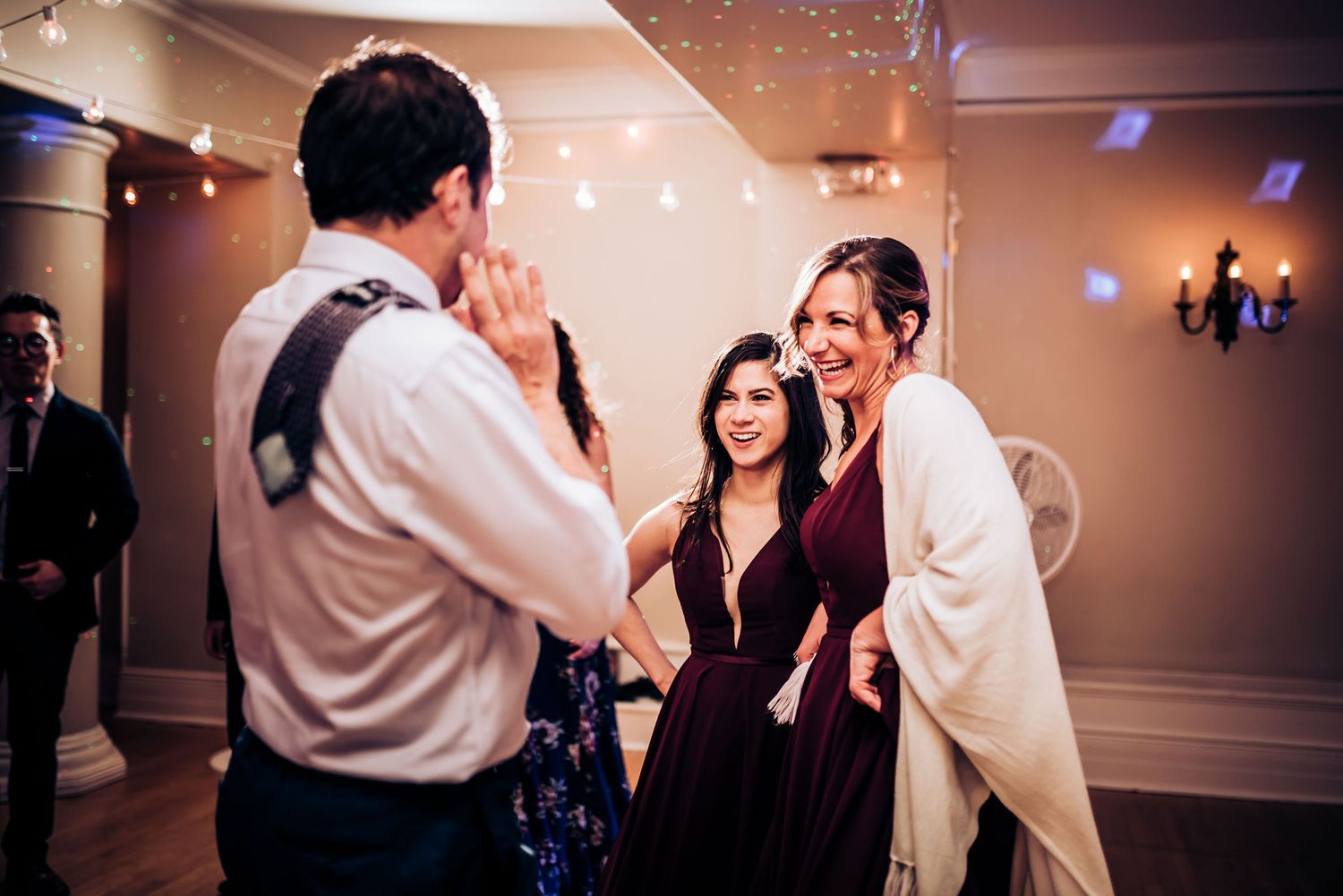 Wedding guests laughing and dancing together at a reception under twinkling lights.