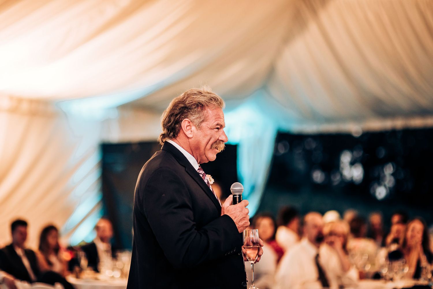 Speaker giving an emotional toast during wedding reception inside elegant tent venue.