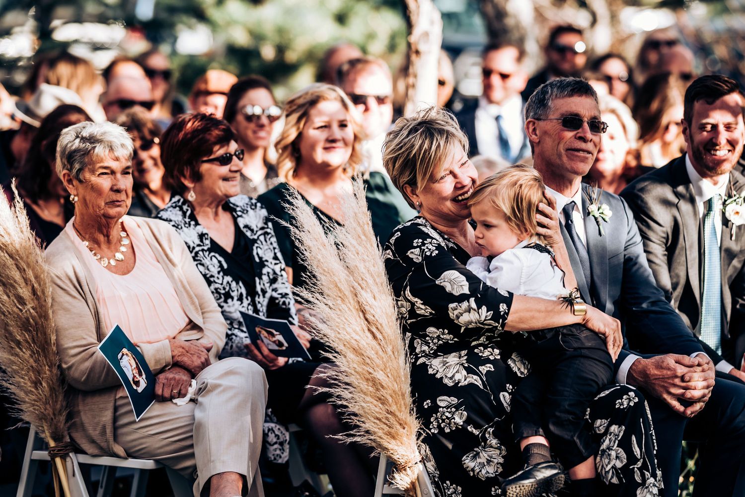 Wedding guests smile and watch the ceremony from white chairs set up on a lawn.