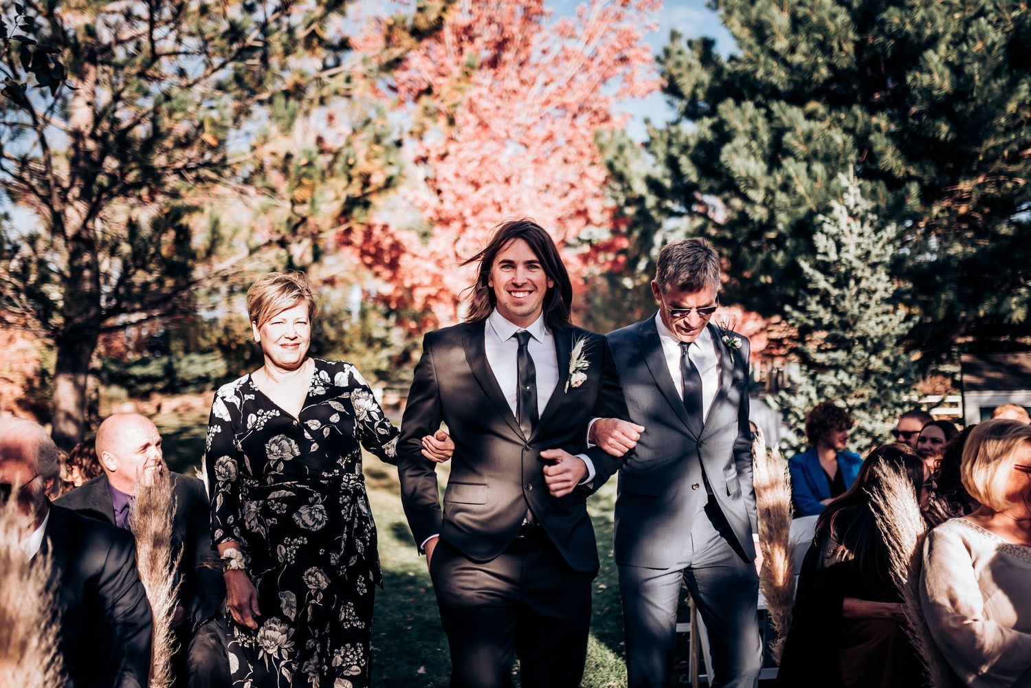 Wedding party members walking down an outdoor aisle surrounded by autumn foliage.