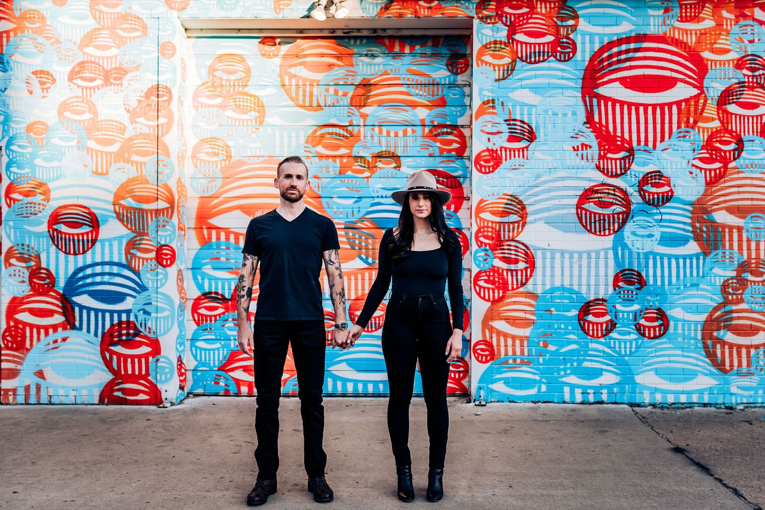 A couple stands holding hands against a vibrant wall mural featuring red and blue circular patterns.