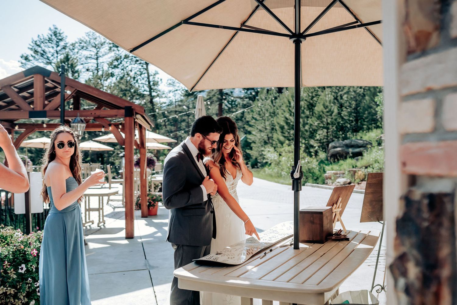 Wedding reception guests gather under cream umbrella at outdoor venue with wooden pergola.