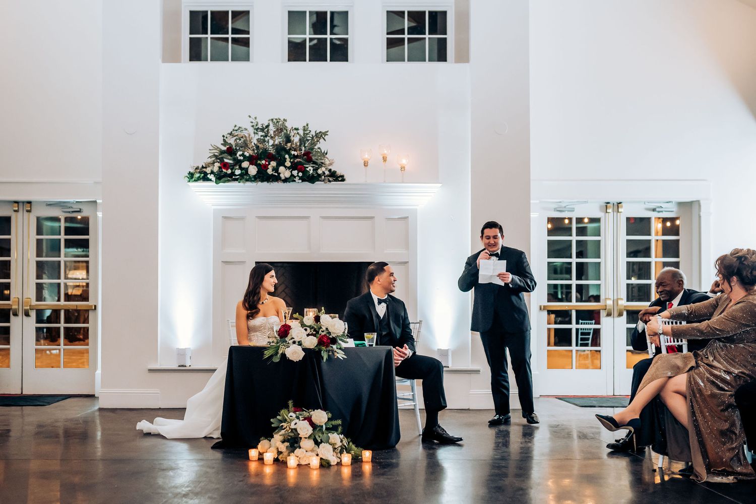 Wedding reception in an elegant white room with fireplace and French doors as guests enjoy speeches and toasts.