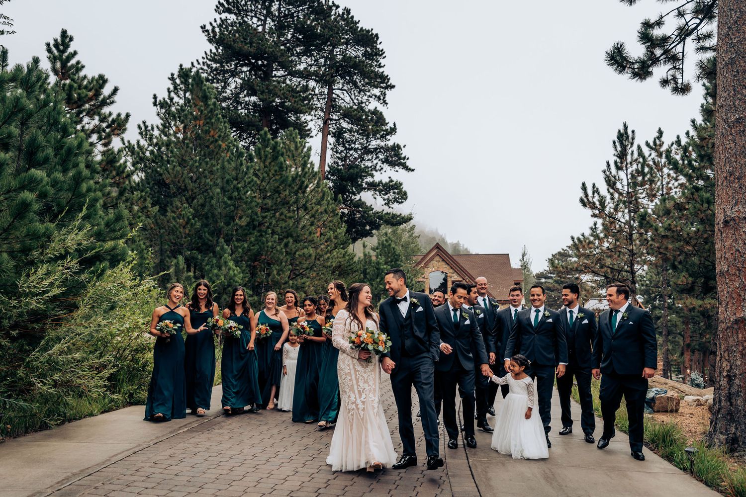Wedding party in teal and navy attire poses on outdoor path with rustic venue in background.
