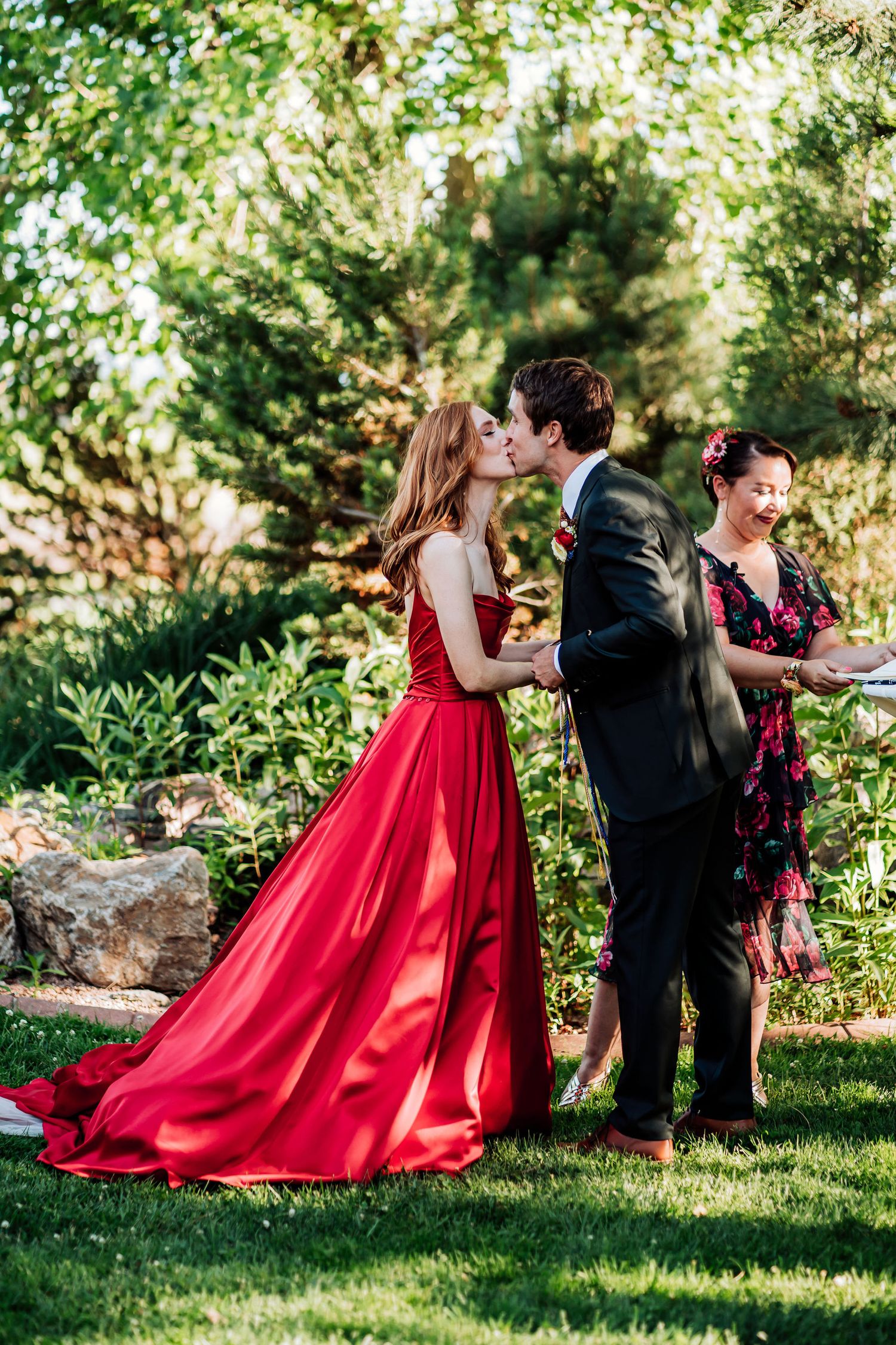 A couple shares a romantic moment on a sunny lawn, with her wearing a flowing red gown and him in a dark suit.