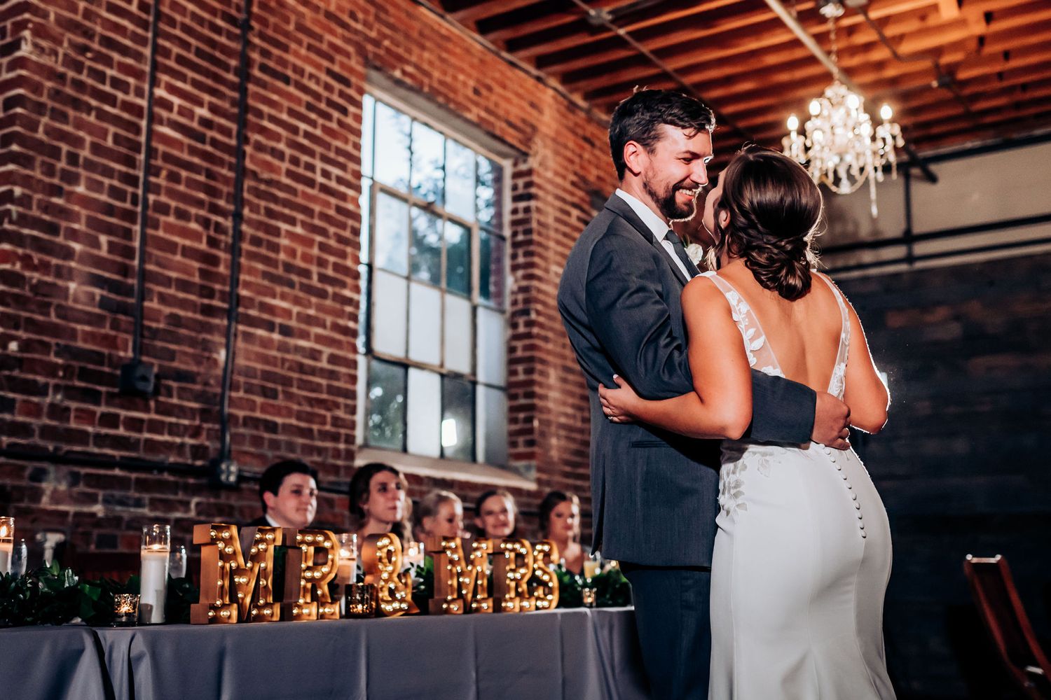 Newlyweds share an intimate dance moment in front of decorated wedding tables and exposed brick walls.