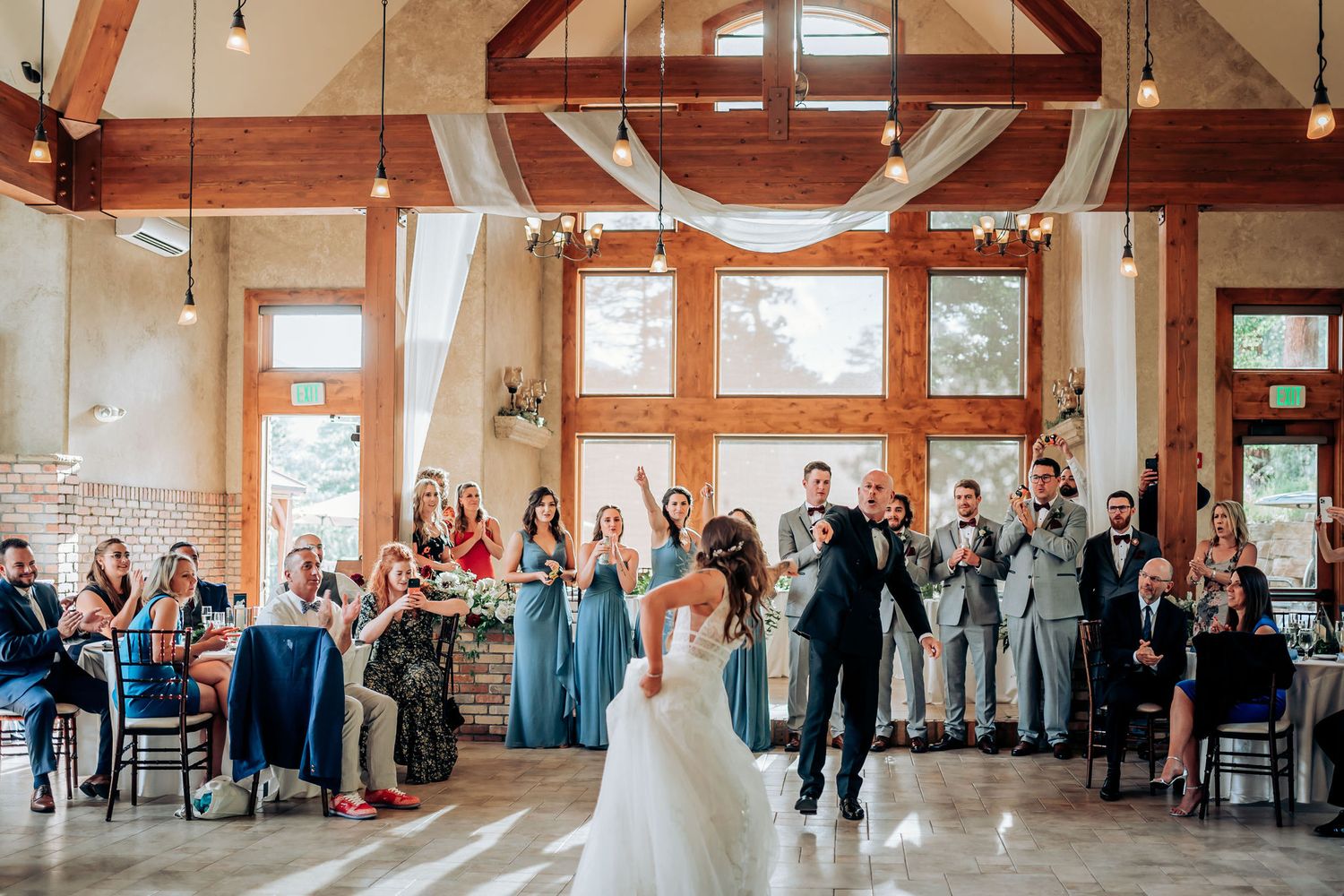 Wedding guests celebrate in a sunlit rustic venue with exposed wooden beams and large windows.