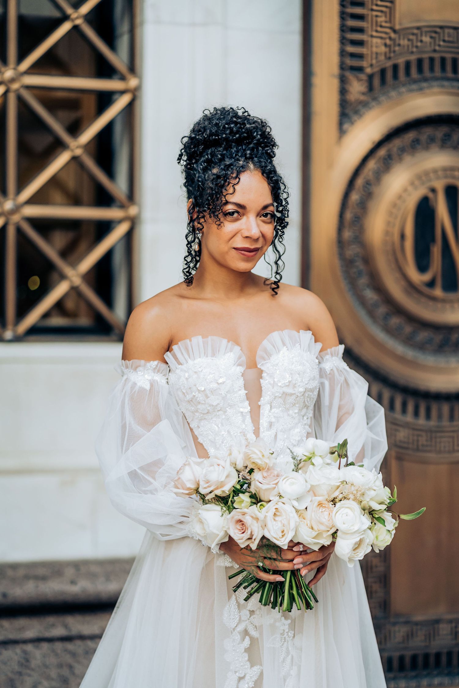 A bride in a strapless white gown with puff sleeves holds a bouquet of pink and white roses.