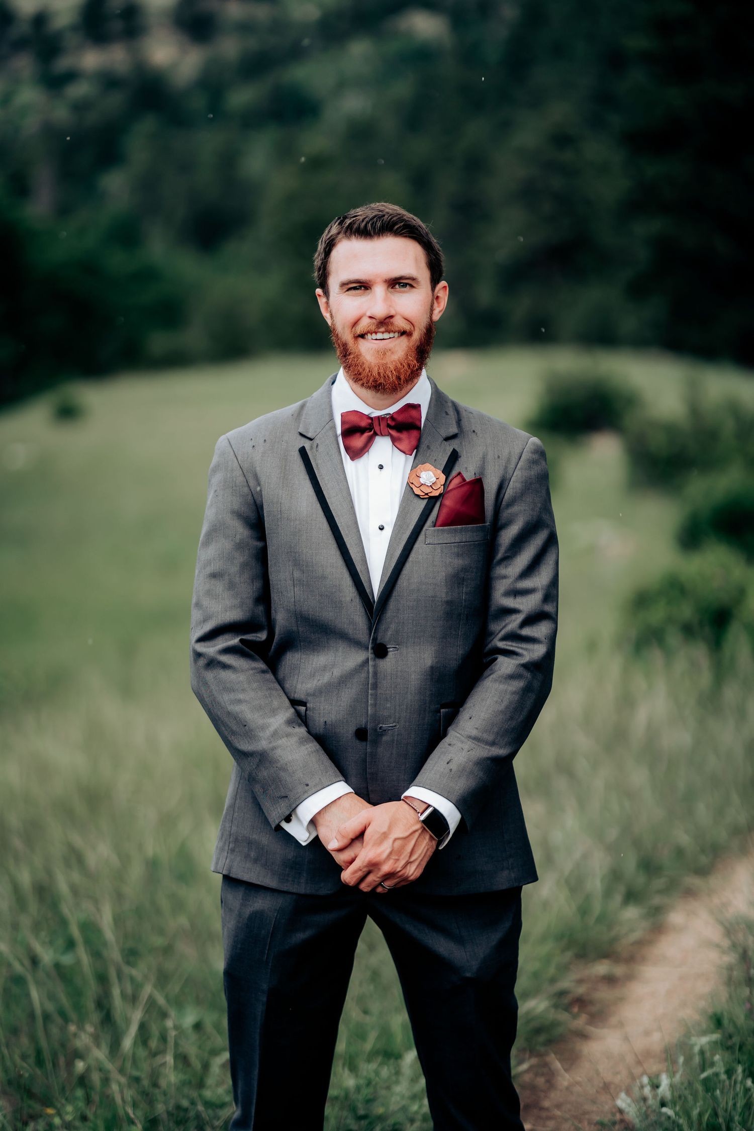 Groom in formal grey suit with red bow tie poses in mountain meadow setting.