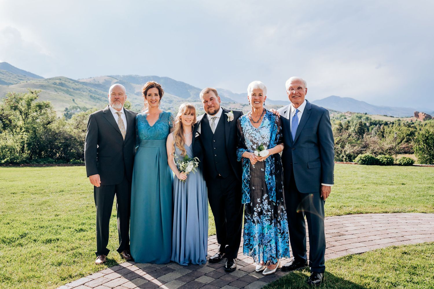 Family group photo on a grassy hillside with mountains in the background during wedding celebration.