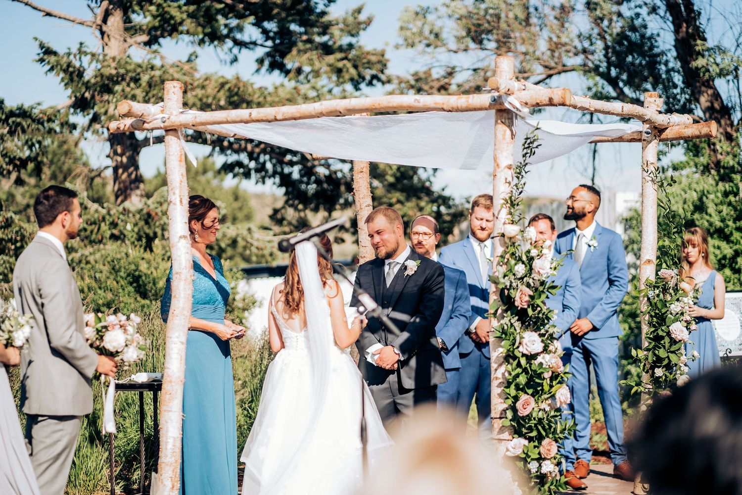 Rustic outdoor wedding ceremony under wooden pergola decorated with white fabric and floral arrangements.