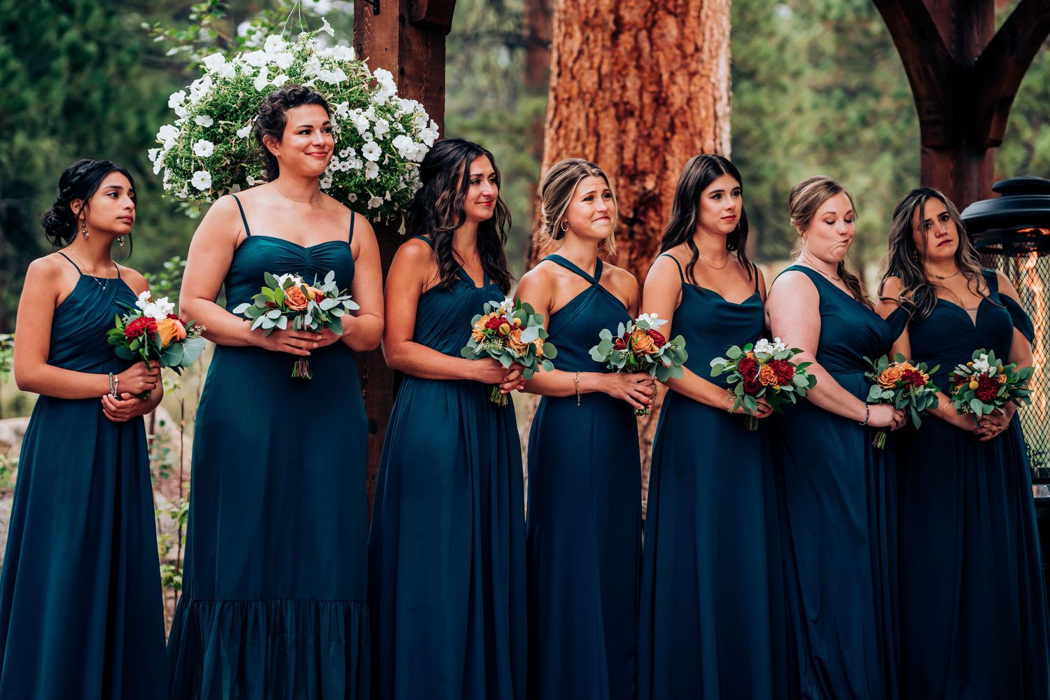 Row of bridesmaids wearing elegant navy blue dresses holding colorful bouquets at an outdoor wedding ceremony.