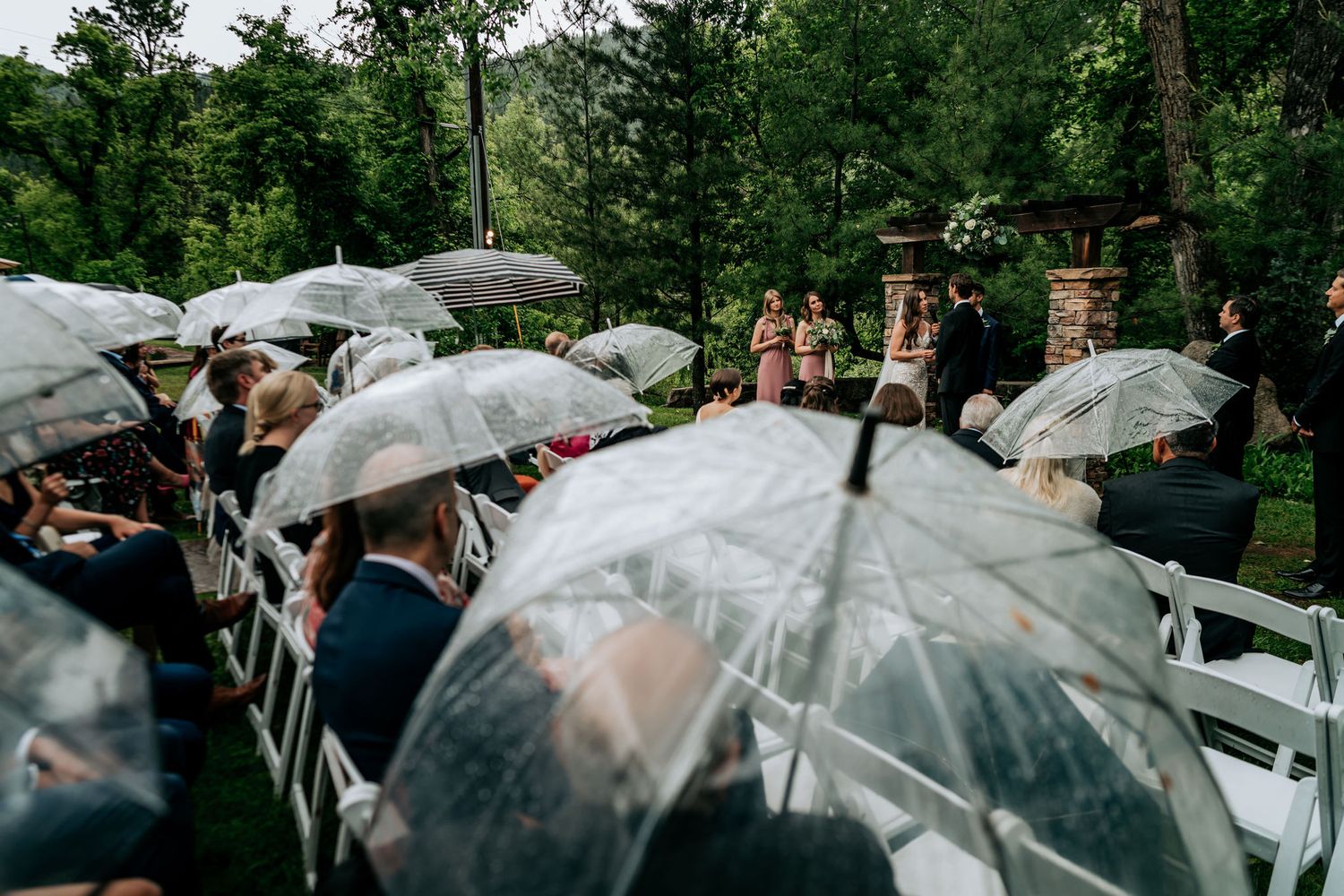 Wedding guests hold clear umbrellas during an outdoor ceremony surrounded by trees.