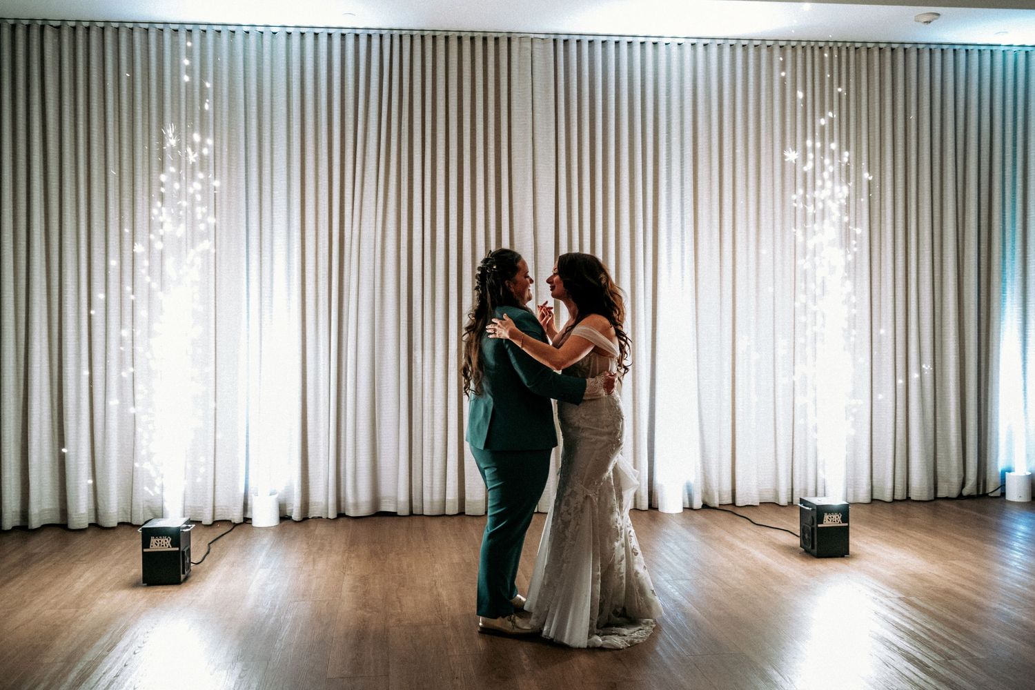 Two brides slow dancing intimately with sparkling light effects against elegant white floor-length curtains.