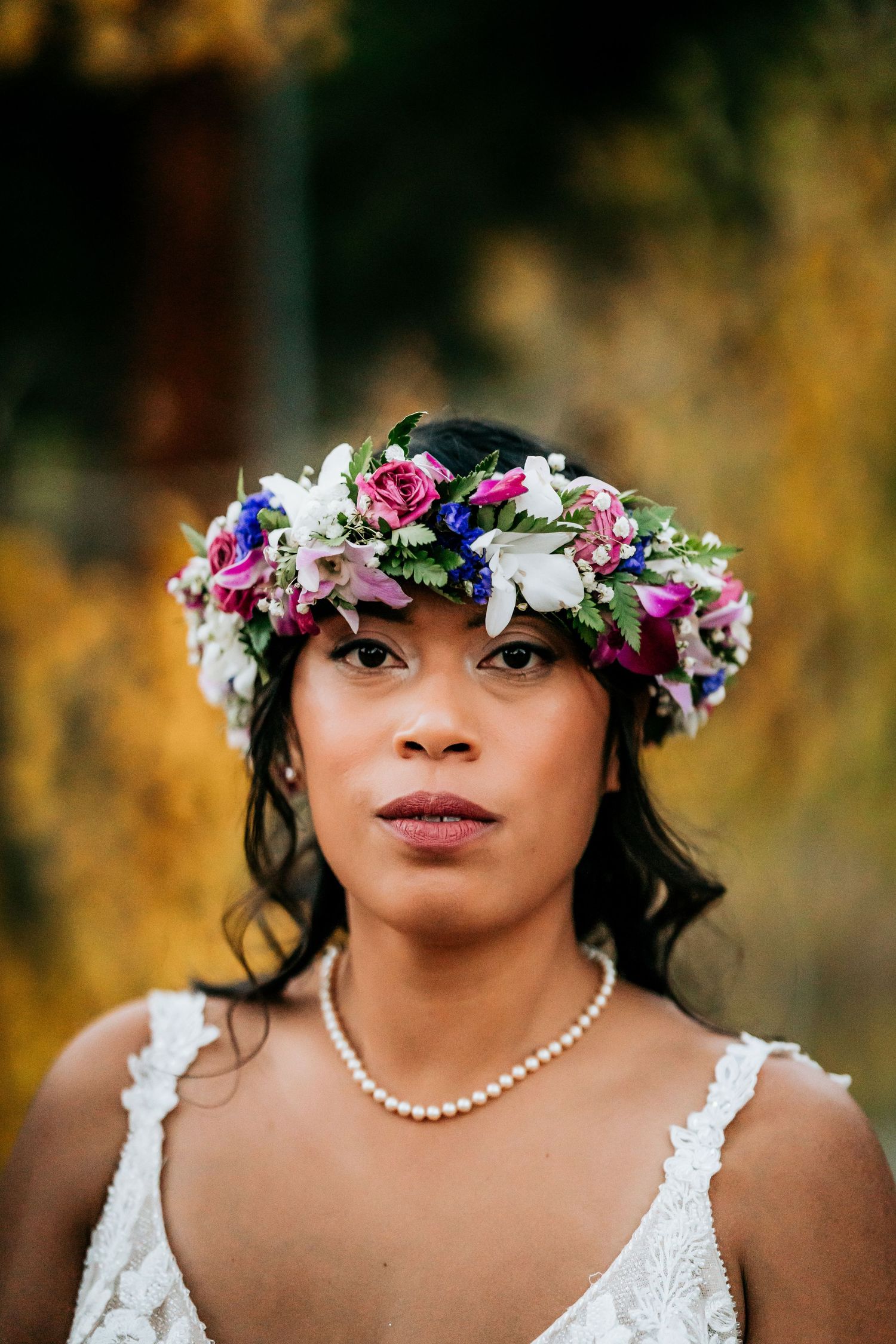 Beautiful floral crown with pink and purple blooms adorns a bride wearing a white lace dress and pearl necklace.
