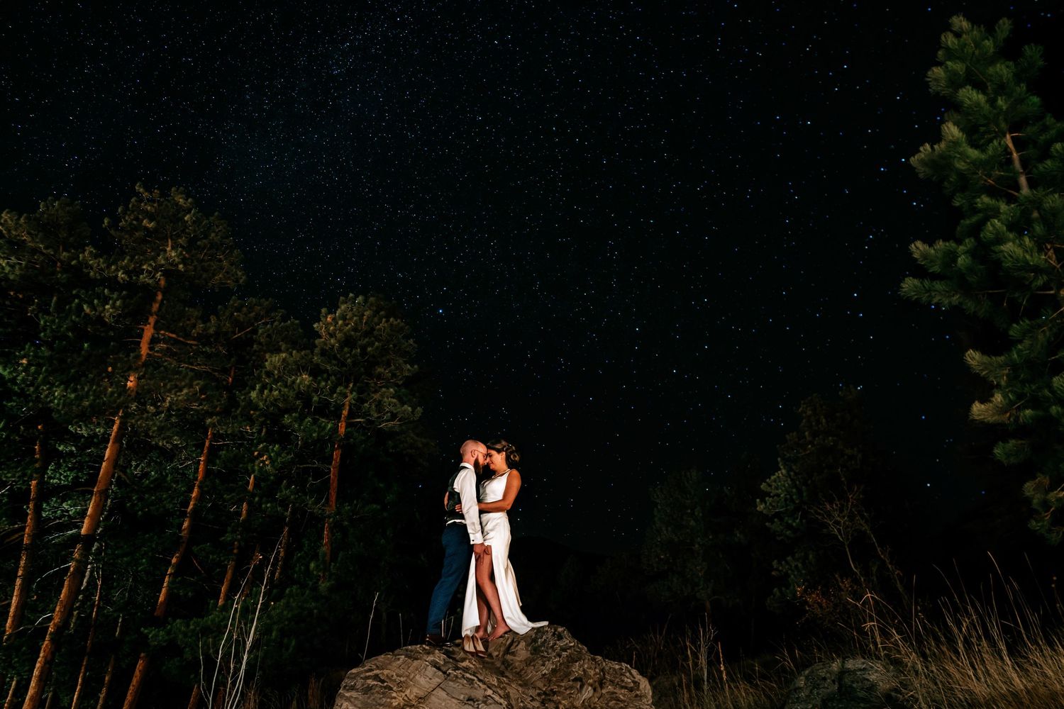A couple shares a romantic moment on a hilltop against a dark sky with pine trees in the background.