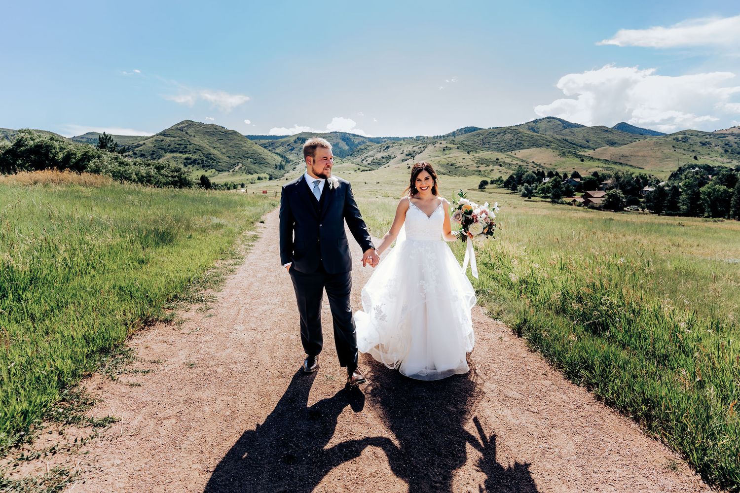Wedding couple walking hand in hand down rural dirt path with mountains in background.