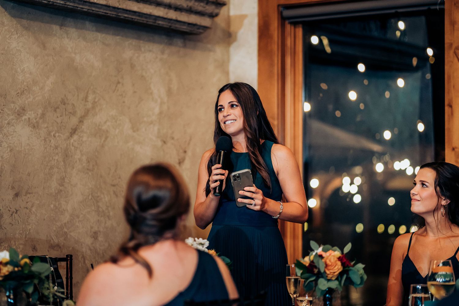 A guest in a navy blue dress gives a speech during an evening wedding reception.