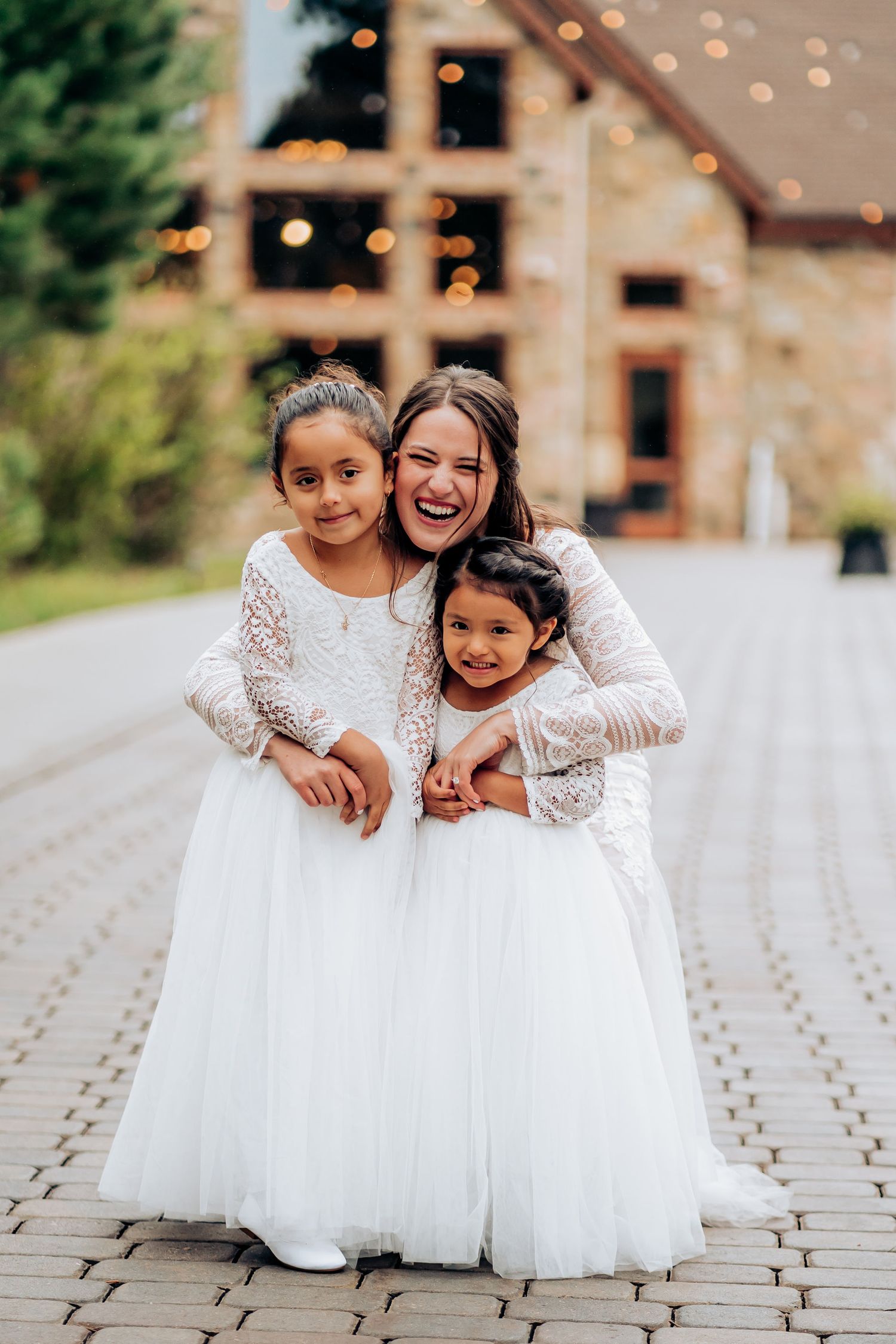 Two flower girls in white dresses share joyful moment with wedding attendant on stone pathway.