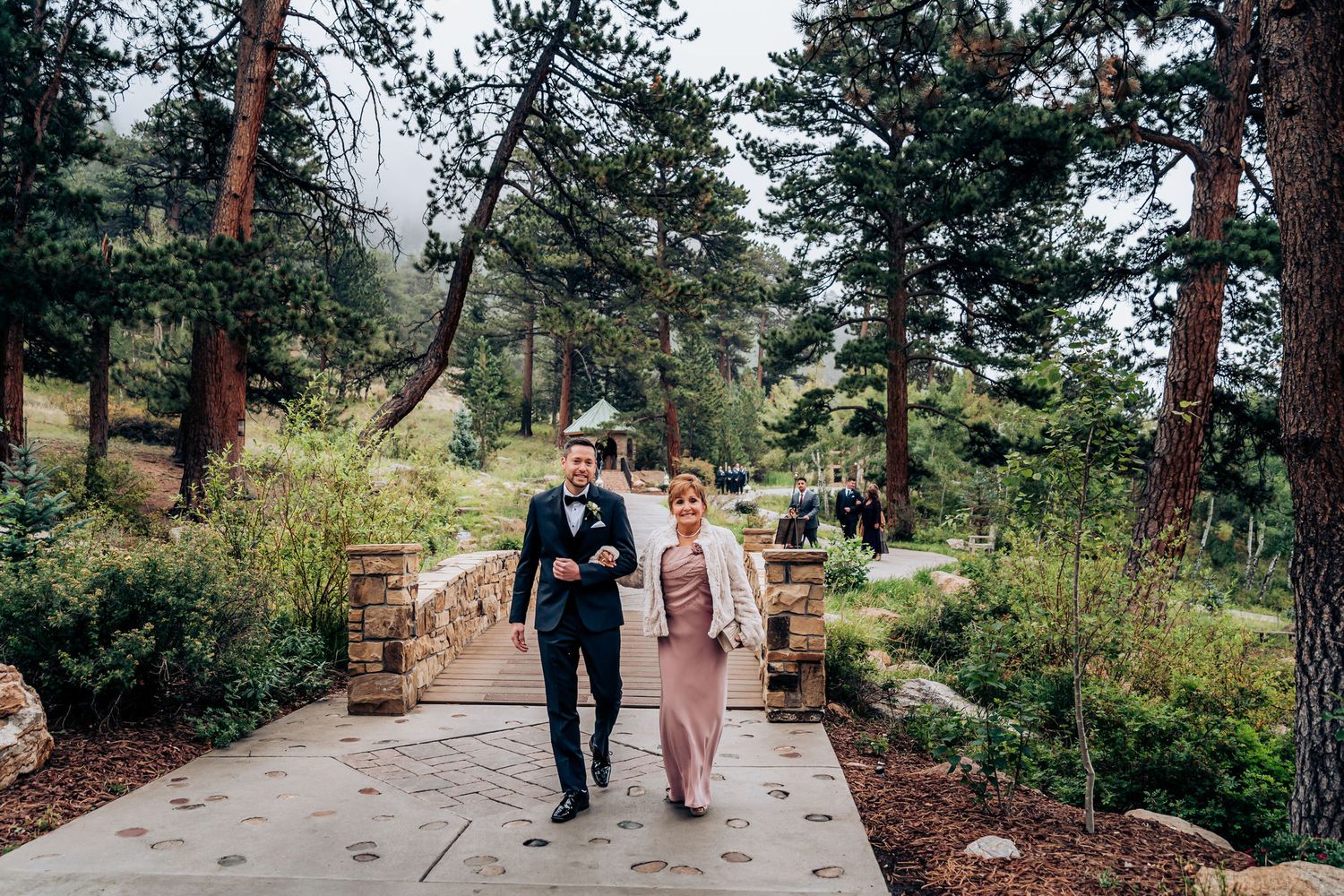 Couple strolling along landscaped pathway with pine trees in mountain setting.