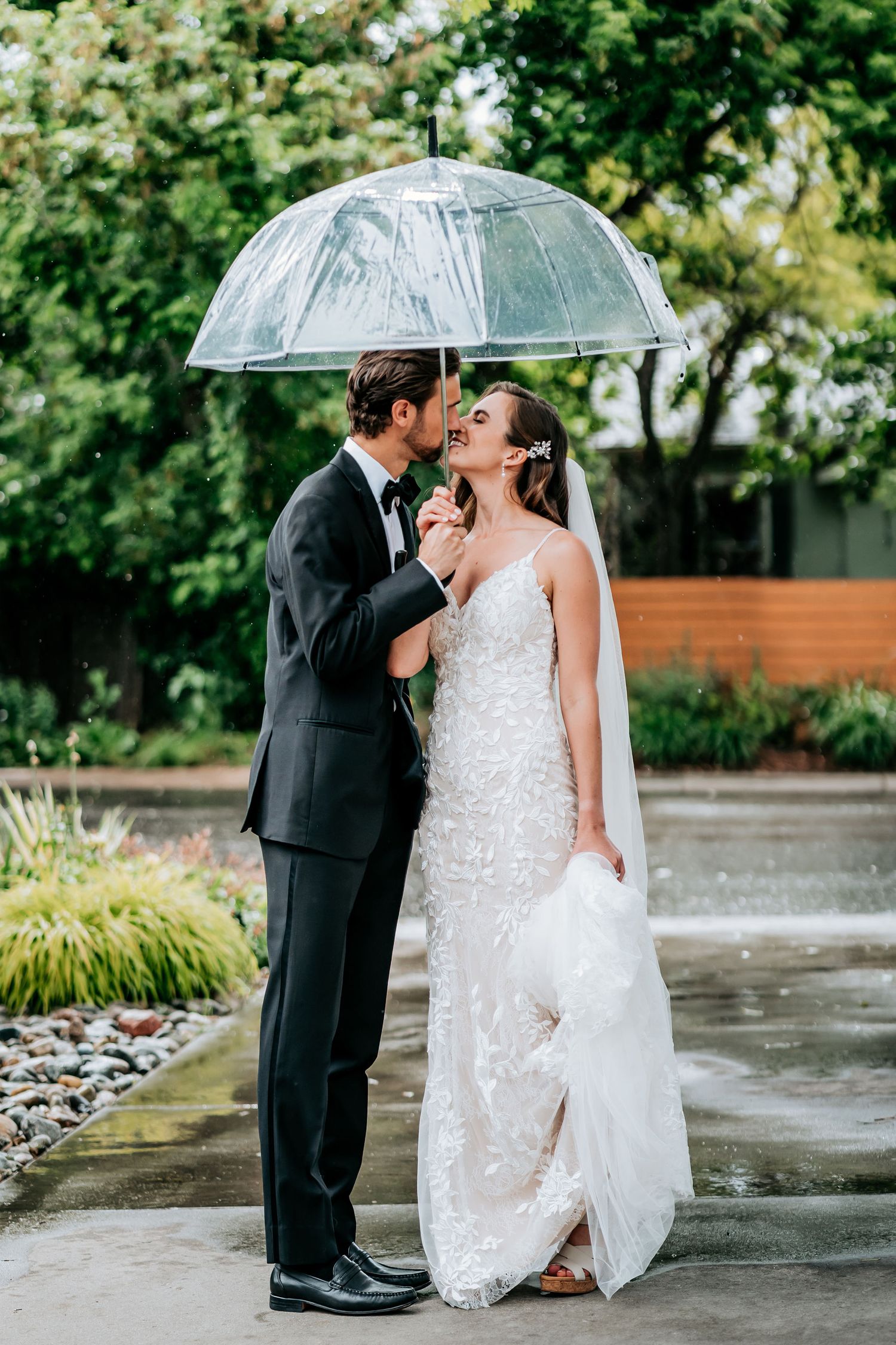 Romantic rainy day wedding portrait under clear umbrella on wet pavement.