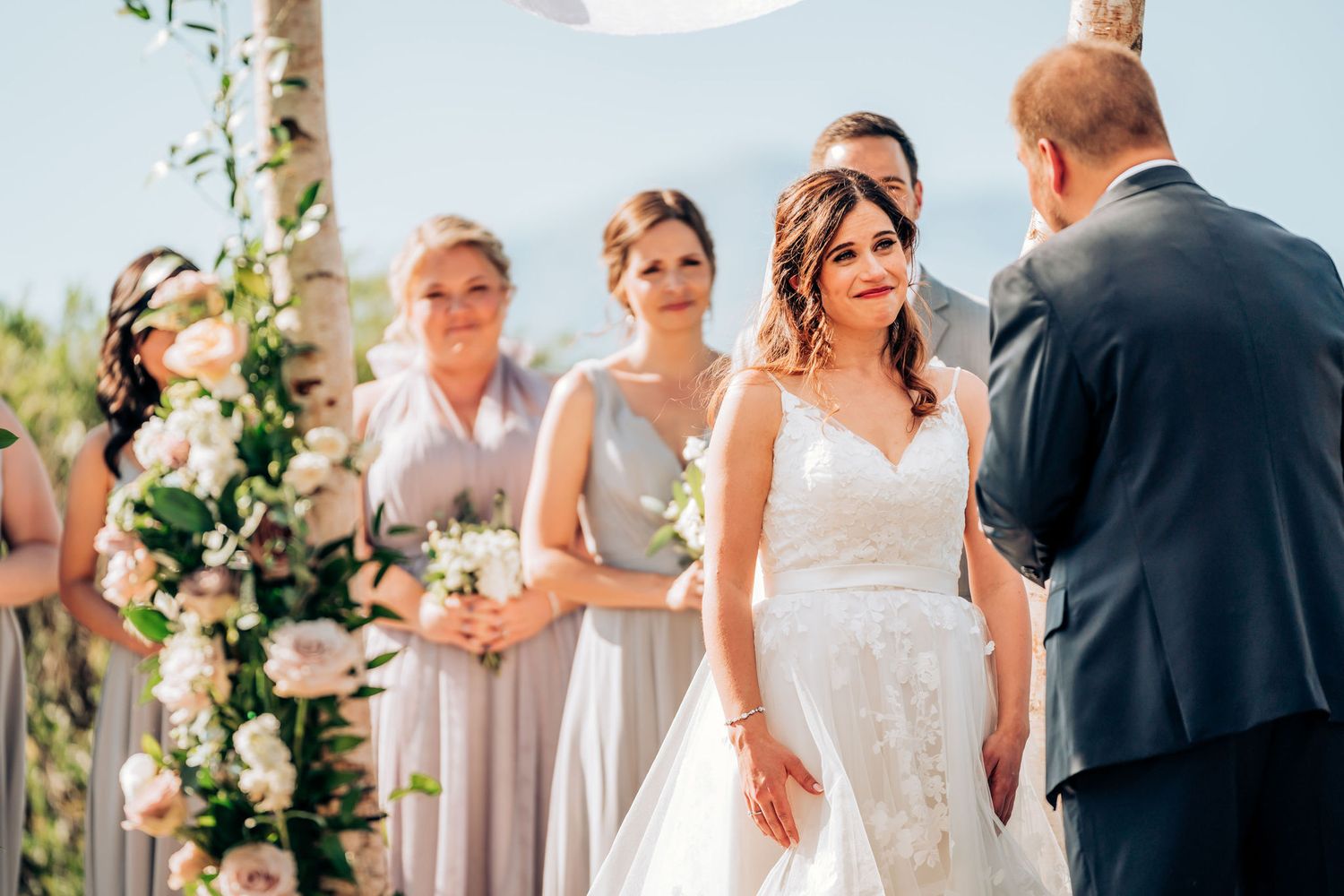 Outdoor wedding ceremony with bridal party standing under floral-decorated birch arch.
